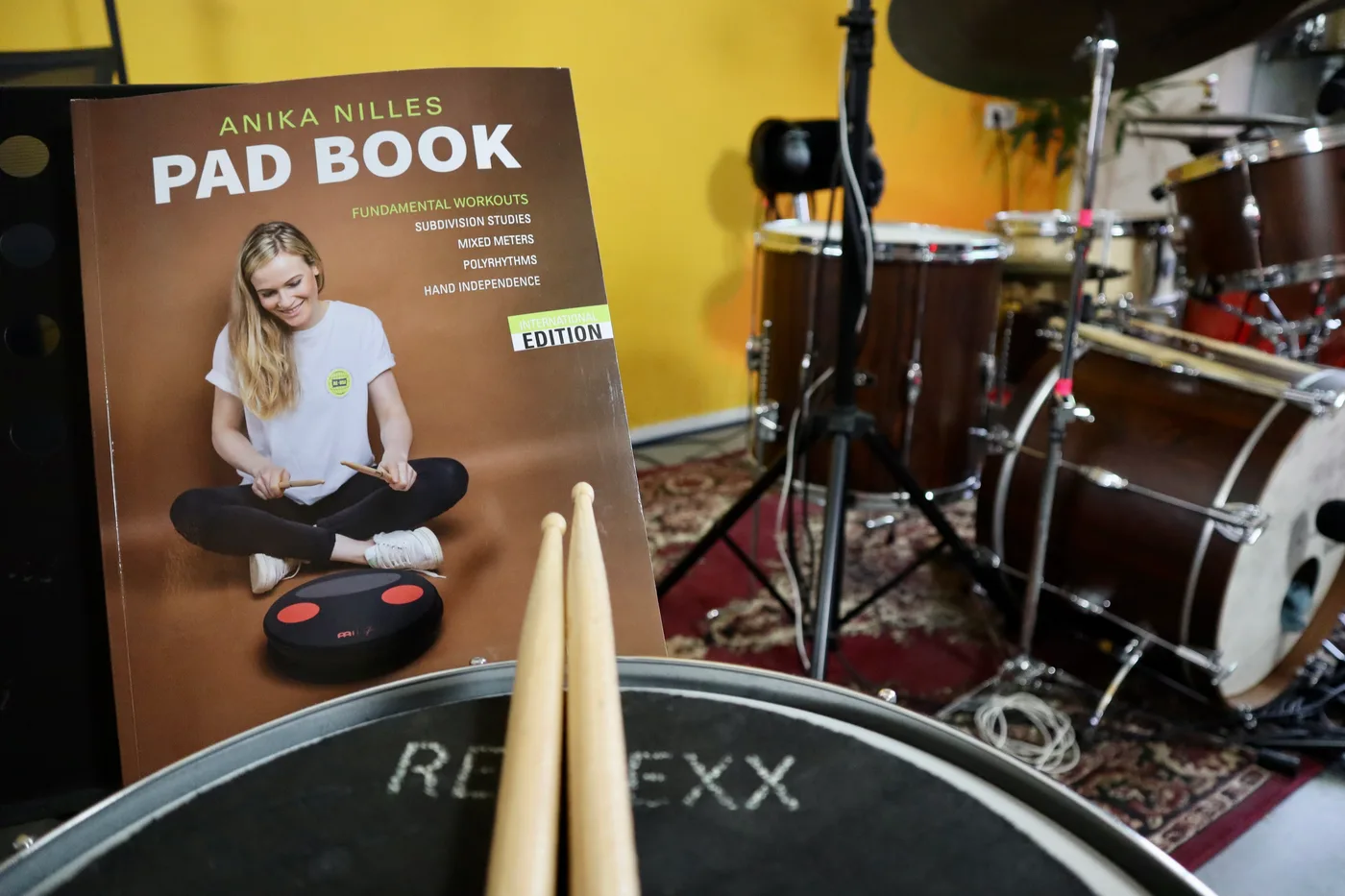 Oefenpad met drumstokken en het Pad Book van Anika Nilles, met het drumstel op de achtergrond
