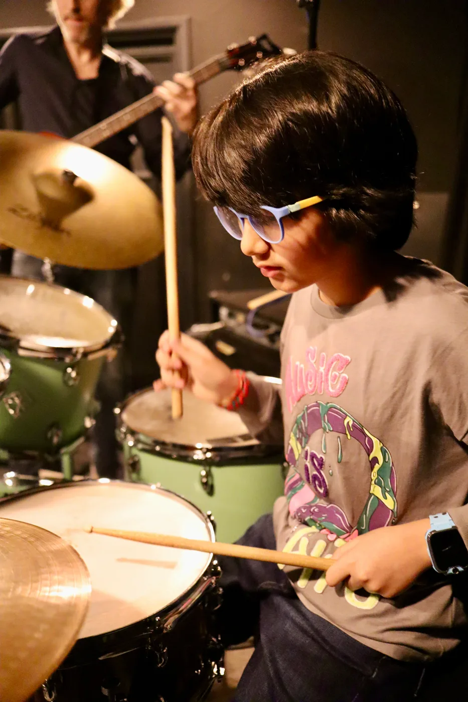 Jongen met bril speelt geconcentreerd drums, met de docent op de achtergrond