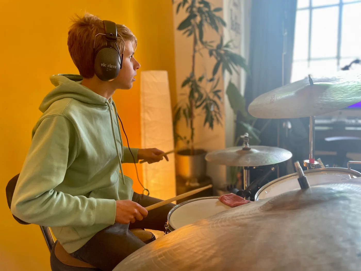 Tiener met koptelefoon en groene hoodie achter het drumstel, voor de gele muur